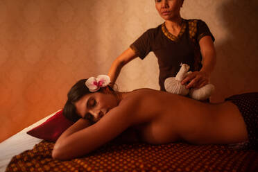 Asian female massagist in traditional clothes giving a thai massage with herbal massage bags to a young attractive woman with orchid on head lying on a bed stretcher in a room lit with warm lights - ADSF40811