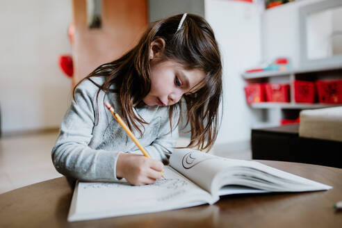Focused girl drawing with pencil in album while sitting at wooden table in light room on blurred background at home - ADSF40822