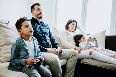 Konzentrierter junger ethnischer Vater mit Sohn, der ein Videospiel spielt, während er auf einem bequemen Sofa in der Nähe von Mutter und Tochter sitzt und auf einem Smartphone surft - ADSF40825