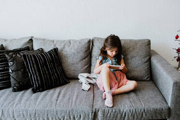 Full body of adorable ethnic little girl in casual clothes and socks relaxing on comfortable sofa and playing video game with smartphone - ADSF40826