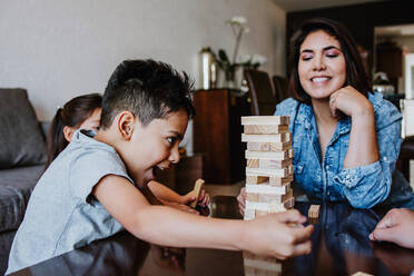 Konzentrierte junge lateinamerikanische Mutter mit Sohn und Tochter, die am Wochenende zu Hause am Tisch sitzend ein Turmspiel spielen - ADSF40827