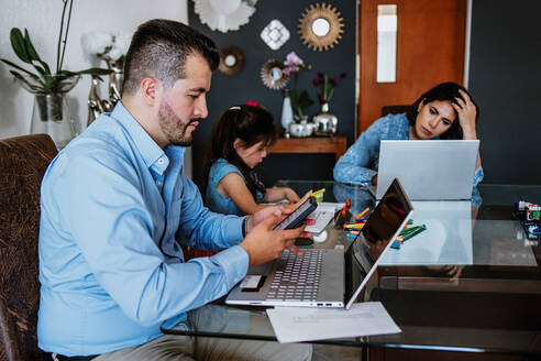Beschäftigte, erschöpfte junge ethnische Eltern, die aus der Ferne an Laptops arbeiten, sitzen zu Hause am Tisch neben ihrer süßen kleinen Tochter, die Hausaufgaben macht - ADSF40831
