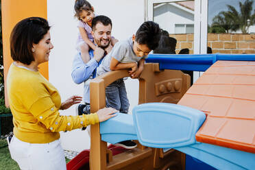 Glückliche lateinamerikanische Mutter und Vater spielen mit niedlichen kleinen Sohn und Tochter auf Rutschen im Hinterhof im Sonnenlicht - ADSF40832