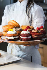 Crop anonymous female baker in uniform standing in bakery with tray of tasty vegan Berliners with whipped cream filling - ADSF40853