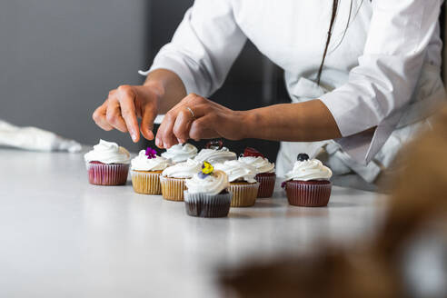 Anonyme Bäckerin dekoriert Cupcakes mit Blumen und Beeren, während sie gesunde vegane Desserts in einer Bäckerei zubereitet - ADSF40855