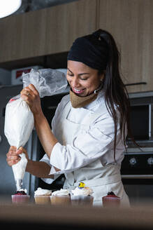 Eine glückliche Bäckerin drückt beim Backen von Desserts in einer Backstube Schlagsahne aus einem Spritzbeutel auf leckere Muffins - ADSF40856