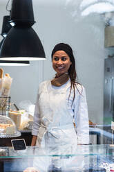 Zufriedene Bäckerin in Uniform steht hinter der Vitrine in der Backstube und schaut weg - ADSF40857