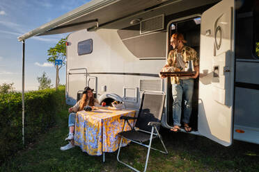 Fröhliches reisendes Paar von Hipstern beim Abendessen am Tisch in der Nähe des Wohnwagens während einer Reise im Sommer in der Natur - ADSF40864