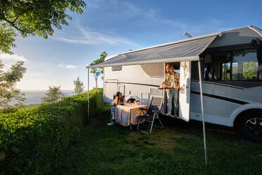 Fröhliches reisendes Paar von Hipstern beim Abendessen am Tisch in der Nähe des Wohnwagens während einer Reise im Sommer in der Natur - ADSF40865
