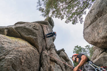 From below climber in helmet holding rope and insuring partner crawling on sheer mountain - ADSF40892