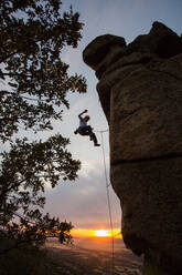 Seitenansicht eines aktiven Bergsteigers mit Schutzhelm, der sich an einem Berghang im warmen Licht des Sonnenuntergangs festhält - ADSF40902