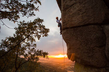 Seitenansicht eines aktiven Bergsteigers mit Schutzhelm, der sich an einem Berghang im warmen Licht des Sonnenuntergangs festhält - ADSF40903
