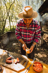 Von oben von ethnischen Mann mit Hut und Vorbereitung leckeren gegrillten Wursthälften auf Brot Stück über Schüssel mit geschnittenen Zwiebeln und Tomaten - ADSF40908