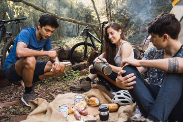Verschiedene Radfahrer sitzen auf dem Boden in der Nähe von Zelt und haben Picknick beim Wandern zusammen im Wald - ADSF40916