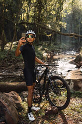 Seitenansicht eines Sportlers mit Schutzhelm, der während eines Ausflugs in den Wäldern mit dem Handy ein Foto von einem Bach macht - ADSF40917