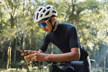 Männlicher Radfahrer mit Helm und Radfahrerbrille telefoniert mit seinem Handy, während er an einem sonnigen Tag im Wald auf dem Fahrrad sitzt - ADSF40918