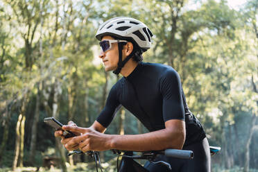 Männlicher Radfahrer mit Helm und Radfahrerbrille telefoniert mit seinem Handy, während er an einem sonnigen Tag im Wald auf dem Fahrrad sitzt - ADSF40919