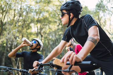 Young sportspeople with water and granola bars having break during bicycle trip in daytime on blurred background - ADSF40921