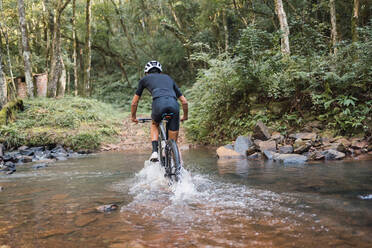 Back view of anonymous male bicyclist riding bike in river with foam during trip in woods - ADSF40922