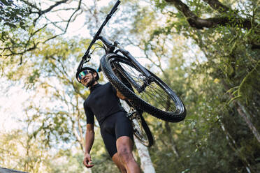 Niedriger Winkel des Sportlers mit Helm und Radfahrerbrille, der sein Fahrrad auf der Schulter trägt, während er auf einem umgestürzten Baumstamm im Wald läuft - ADSF40923