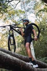 Niedriger Winkel des Sportlers mit Helm und Radfahrerbrille, der sein Fahrrad auf der Schulter trägt, während er auf einem umgestürzten Baumstamm im Wald läuft - ADSF40924