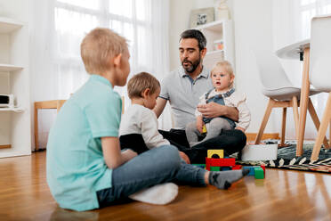 Dad speaking with children while playing with soft blocks on parquet on weekend at home - ADSF40930