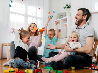 Cheerful parents with children throwing module blocks into air while playing in light house room - ADSF40932