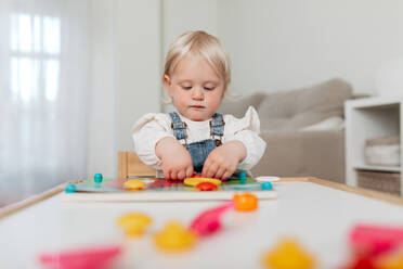 Charming little kid with blond hair assembling game pieces at table at home - ADSF40935