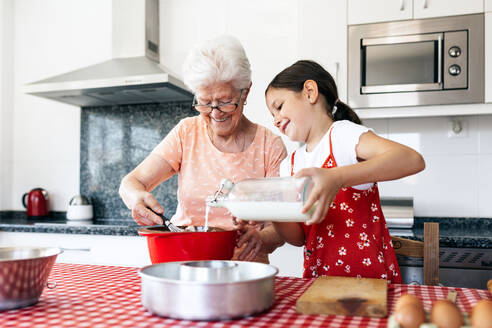 Großmutter mit Brille und Schneebesen in der Nähe der Enkelin mit Milchflasche beim Teigkochen am Tisch im Haus - ADSF40937