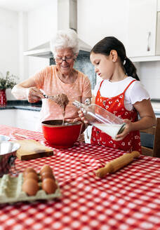 Großmutter mit Brille und Schneebesen in der Nähe der Enkelin mit Milchflasche beim Teigkochen am Tisch im Haus - ADSF40938