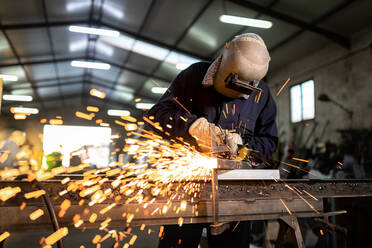Unbekannter männlicher Angestellter mit Schutzhandschuhen und Helm bei der Arbeit an einem Schweißgerät in einer dunklen Werkstatt - ADSF40950