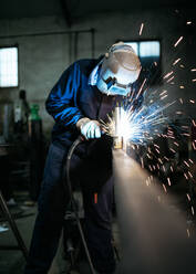 Unbekannter männlicher Angestellter mit Schutzhandschuhen und Helm bei der Arbeit an einem Schweißgerät in einer dunklen Werkstatt - ADSF40952