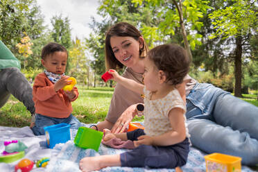 Kleines Baby Mädchen und Junge sitzen auf Plaid mit Spielzeug in der Nähe Ernte Eltern im Park - ADSF40977