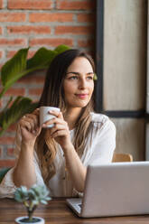 Glückliche junge Frau mit langen Haaren nippt an frischem Kaffee aus einer Tasse, während sie in der Pause einer freiberuflichen Tätigkeit in einer Cafeteria mit ihrem Laptop am Tisch sitzt und wegschaut - ADSF40982