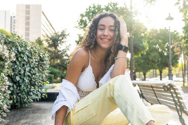 Fröhliche junge hispanische Frau in stilvoller Kleidung, die auf einer Marmorplatte sitzt und lächelnd in die Kamera blickt, während sie auf einer Marmorplatte in der Nähe von üppigen Büschen an einem sonnigen Sommertag im Park sitzt - ADSF40983