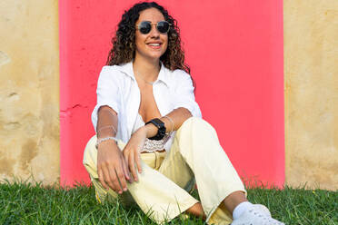 Voller Körper froh junge hispanische Frau in stilvolle Kleidung und Sonnenbrille lächelnd und Blick auf die Kamera, während auf grasbewachsenen Rasen gegen rosa Wand sitzen - ADSF40984