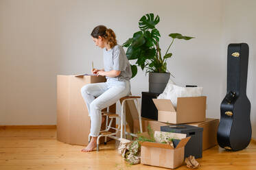 Side view of young female in casual clothes writing down data in notebook on cardboard box while sitting on ladder in light room of new flat - ADSF40987