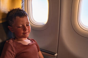 High angle of calm boy smiling while sitting on chair and sleeping near windows in plane - ADSF40992