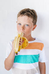 Adorable kleiner Junge mit braunen Haaren in T-Shirt lächelnd und Blick auf die Kamera beim Essen leckere Banane gegen weißen Hintergrund - ADSF40998