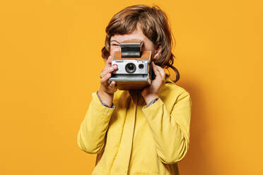 Kleines Mädchen in leuchtend gelber Herbstjacke, das ein Foto mit einer Vintage-Kamera vor einem gelben Hintergrund im Studio aufnimmt - ADSF41004