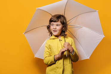 Kleines Mädchen in leuchtend gelber Herbst-Kapuzenjacke und Regenschirm schaut weg vor gelbem Hintergrund im Studio - ADSF41005