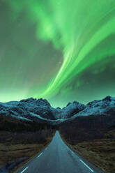 Landschaftlicher Blick auf ein Auto, das auf einer leeren Bergstraße im Winter fährt, während der Nachthimmel mit hellen grünen Polarlichtern in Norwegen leuchtet - ADSF41010