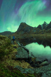Unrecognizable traveling photographer taking picture of green aurora borealis shining in night sky in Lofoten Islands near pond in rocky snowy mountains - ADSF41016