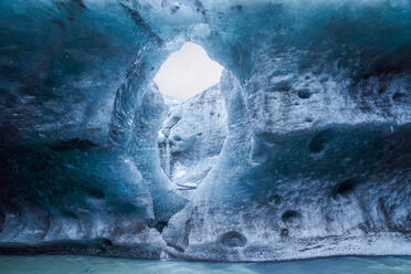 Unebenes Loch in der Eiswand, das Sonnenlicht in eine Höhle mit einem Strom aus kaltem Wasser im Vatnajokull-Gletscher an einem Wintertag in Island lässt - ADSF41065