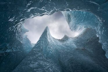 Von unten Eisspitzen, die das Loch in der Höhlendecke umgeben, gegen einen bedeckten grauen Himmel an einem Wintertag im Vatnajokull-Nationalpark in Island - ADSF41066