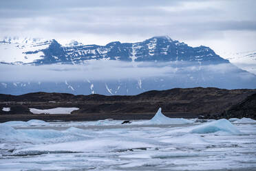 Landschaft mit Eisstücken, die im Meer schwimmen, umgeben von schneebedeckten Bergen im vulkanischen Gebiet des Vatnajokull-Nationalparks an einem bewölkten Tag in Island - ADSF41080