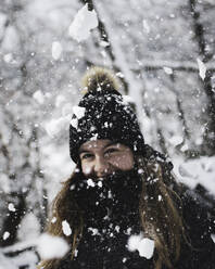 Fröhliche junge Touristin mit langen braunen Haaren in warmer Oberbekleidung und Strickmütze, die fröhlich lächelt und in die Kamera schaut, im verschneiten Wald an einem Wintertag - ADSF41084