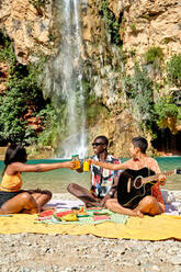 Group of multiracial tourists sitting on beach against lake and clinking bottles of juice while enjoying summer vacation together - ADSF41102