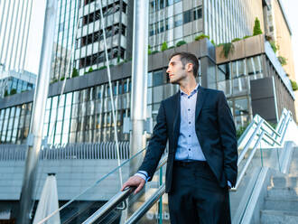 Low angle of pondering entrepreneur standing on steps in front of modern corporate building and looking away with hand in pocket - ADSF41119