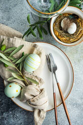 From above cutlery set for Easter dinner with olive tree branches and eggs on a bird nest on a concrete background - ADSF41123